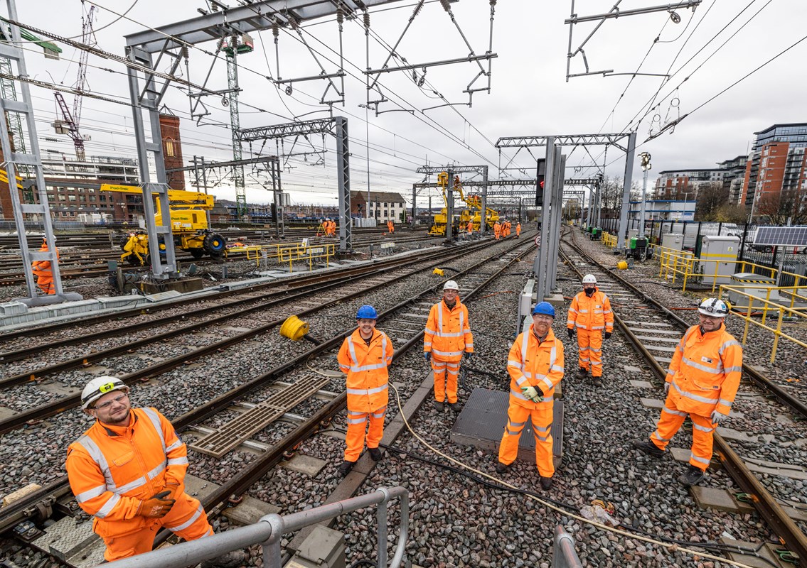 Rail engineering crew in hi-vis on track amid overhead line infrastructure.